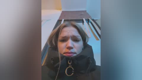 Media: Video of a young Caucasian woman with shoulder-length brown hair, wearing a black jacket, restrained with a gag and handcuffs, in a room with white walls and wooden planks on the floor.