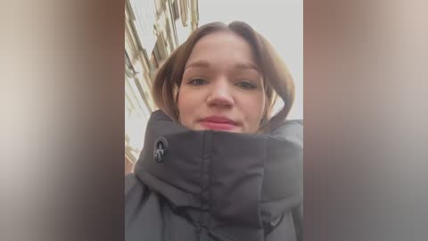 Media: Video of a woman with light skin and shoulder-length brown hair, wearing a black puffy jacket, looking directly at the camera. The background shows a blurred, beige building.