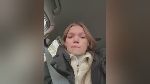 Media: Video of a young woman with light skin, shoulder-length brown hair, wearing a black jacket and beige sweater, seated in a car, with blurred background.