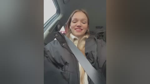 Media: A video captures a young woman with fair skin, blonde hair, and closed eyes, smiling, wearing a black puffy jacket and beige scarf, sitting in a car with a seatbelt across her chest.