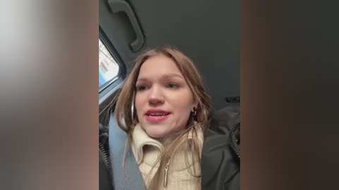 Media: Video of a young Caucasian woman with light brown hair, wearing a beige sweater and black jacket, smiling inside a bus with a window and overhead compartment visible.