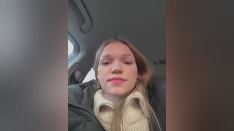 Media: A video of a young woman with fair skin and long, straight, light brown hair, wearing a dark green jacket and beige sweater, sitting in a car. The background shows a gray interior and a window.