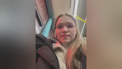 Media: Video of a blonde woman in a subway car, wearing a black jacket over a beige sweater, looking straight ahead.