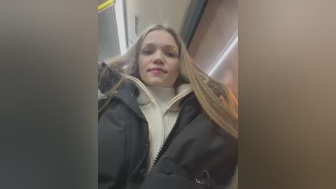 Media: Video of a young woman with long blonde hair, wearing a black puffer jacket over a cream-colored sweater, standing in a subway car.