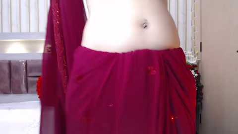 Media: Video of a fair-skinned person, midsection visible, draped in a red, semi-transparent sari with a subtle floral design. Background features a white door and a beige wall.