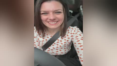 Media: A video of a smiling woman with shoulder-length dark hair, wearing a white shirt with red star pattern, seated in a car with a safety belt.