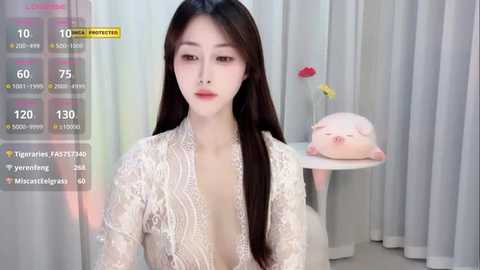 Media: Video of an Asian woman with long black hair, wearing a sheer, white lace dress, sitting in a room with white curtains and a pink plush toy on a table.