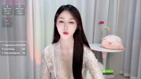 Media: Video of a fair-skinned East Asian woman with long black hair, wearing a white lace robe, sitting in front of a white table with a pink piggy bank and flowers.