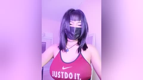 Video of a woman with medium skin tone, wearing a red Nike sports bra with \"JUST DO IT\" in white, a black mask, and black hair in a high ponytail. She stands in a softly lit room with pastel walls.