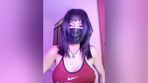 Video of an Asian woman with straight black hair, wearing a black face mask, red sports bra, and choker necklace, standing in a dimly lit room.