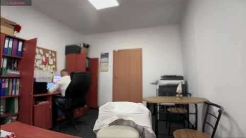 Media: Video of a small, cluttered office with white walls, a wooden desk, black chair, bookshelf, printer, and two men in shirts and pants.