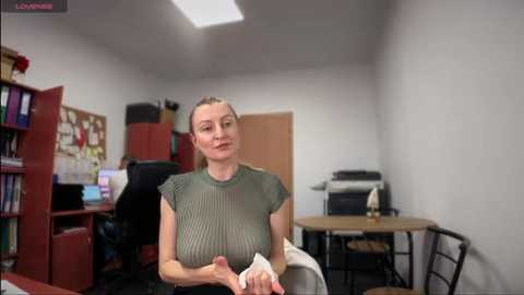 Media: Video of a blonde woman with a slender build, wearing a green mesh top, sitting in a sparsely furnished office with a wooden desk and bookshelf.