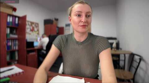 Video of a blonde woman with light skin in a grey ribbed top, sitting at a desk in a classroom. Background shows shelves with books, a bulletin board, and chairs.