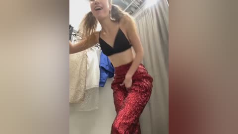 Media: Video of a light-skinned woman with long, light brown hair in pigtails, wearing a black sports bra and red sequin pants, laughing in a brightly lit dressing room with hanging clothes.