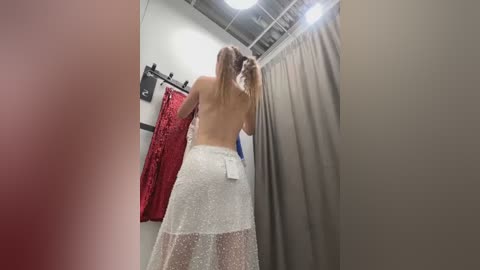 Media: Video of a topless woman with long blonde hair in a changing room, wearing a sheer, glittery white skirt. Red and beige curtains in the background, fluorescent light overhead.