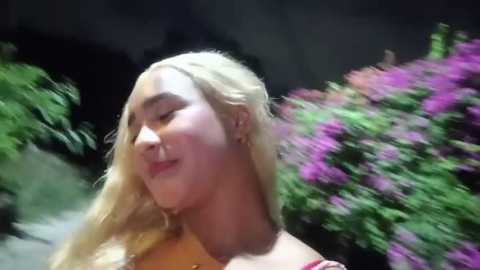 Media: A video of a young, fair-skinned woman with long, blonde hair, smiling gently, wearing a pink top. The background is blurred, featuring lush greenery and vibrant purple flowers.