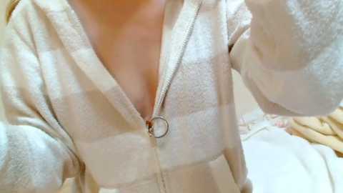 Media: A close-up video of a person wearing a white and beige plaid hoodie with a zipper and silver ring pull, their skin visible through the hoodie. The background is blurred, suggesting an indoor setting.