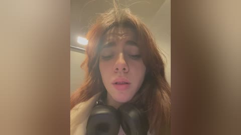 Media: Video of a young woman with long, reddish-brown hair, wearing black headphones, eyes closed, head tilted back, looking serene in a dimly lit room with a beige ceiling.