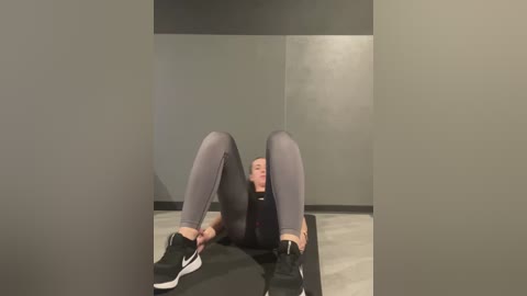 Media: Video of a white woman with short blonde hair, wearing tight grey leggings and black Nike sneakers, sitting on a yoga mat in a minimalist room with grey walls and light wooden floor.