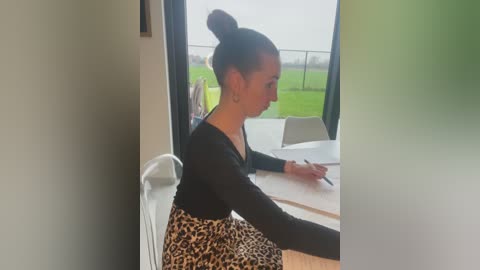 Media: Video of a fair-skinned woman with dark hair in a bun, wearing a black long-sleeve top and leopard-print skirt, seated at a white table, writing with a pen. Background shows a green field and a fence.