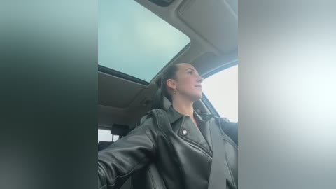 Media: Video of a woman in a black leather jacket, driving a car with a light gray interior. She has short brown hair and is looking to the right.