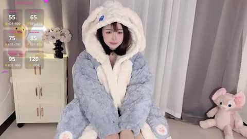 Media: Video of an East Asian woman with medium skin tone and long black hair, wearing a blue plush robe, seated on a cream-colored floor, in a minimalist room with gray curtains and a pink stuffed bear.