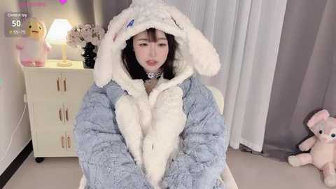 Media: Video of an Asian woman in a fluffy, light blue bunny costume, sitting on a bed with white curtains. She has fair skin and short dark hair. A stuffed bunny is on the floor, and a nightstand with a lamp and flowers is visible.