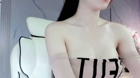 Media: Video of an Asian woman with pale skin and long black hair, wearing a strapless beige top with bold black text, seated in a white, cushioned chair in a dimly lit, marble-floored room.