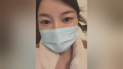 Media: Video of an Asian woman with fair skin and dark hair, wearing a light blue surgical mask, and a white bathrobe.