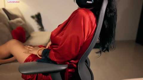 Media: A video showing a woman with long black hair and fair skin, wearing a red satin robe, seated in a grey gaming chair, with a man partially visible on a couch in the background.