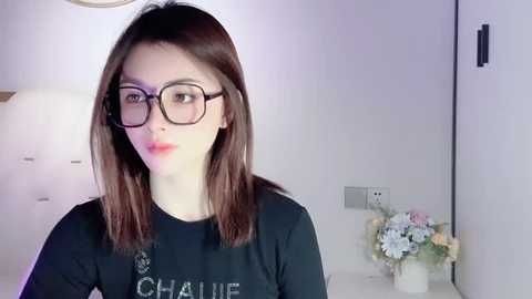 Media: Video of a young woman with straight, shoulder-length brown hair, wearing black-rimmed glasses and a black \"CHANEL\" t-shirt, standing in a modern, white-walled room with a vase of flowers on a white table.