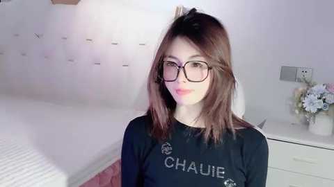 Media: A video of a young woman with fair skin, shoulder-length brown hair, and glasses, wearing a black \"CHANEL\" sweatshirt, standing in a modern bedroom with a white bed, nightstand, and flowers.