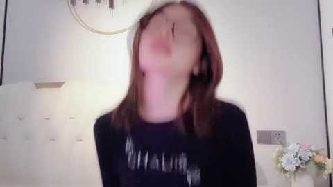 Media: A video of a young woman with straight, shoulder-length brown hair, wearing a black shirt, captured mid-laugh, with a blurred background of white walls and a small flower arrangement.