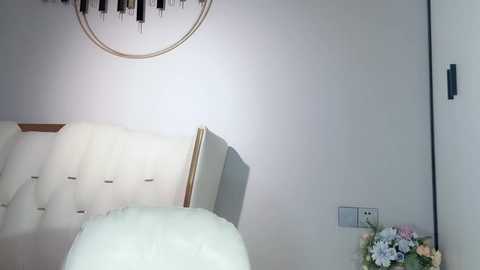 Media: Video of a minimalist bedroom with a tufted white headboard, white duvet, light gray walls, circular pendant light, and a small bouquet of pastel flowers on a side table.