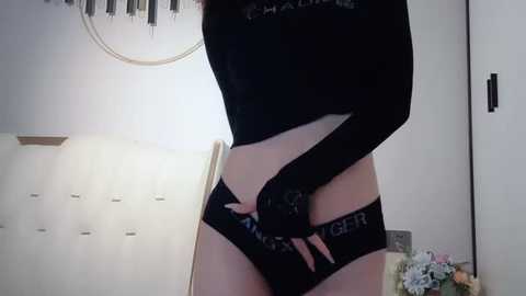 Media: Video of a slender, light-skinned person with long, dark hair, wearing a black crop top and black panties with \"GIRL\" branding, standing in a minimalist room with a wooden chair and white walls.