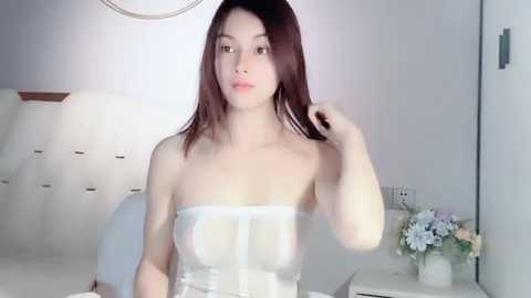 Media: Video of a young Asian woman with long, straight brown hair, wearing a sheer, strapless white dress, standing indoors near a white couch and a floral arrangement.