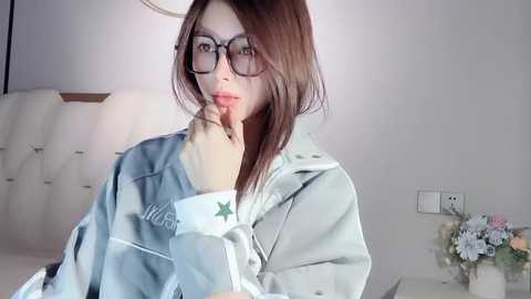 Video of a young East Asian woman with straight brown hair, wearing glasses and a light gray hoodie, sitting on a bed in a softly lit, minimalistic bedroom.