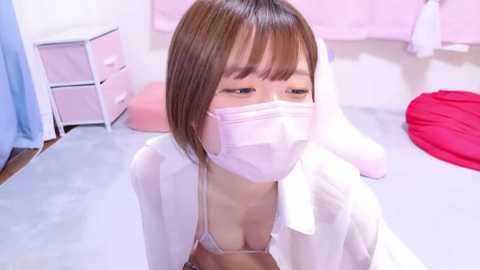 Media: Video of an Asian woman with straight brown hair and light skin, wearing a white mask, light bra, and robe, kneeling on a light gray floor in a pastel-colored room with pink curtains and red beanbag.