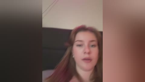 Media: Video of a young woman with long, straight, reddish-brown hair, fair skin, and light makeup, slightly blurred. She has a neutral expression and is indoors, possibly in a bedroom with dark furniture.