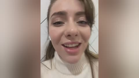 Media: A close-up video of a young woman with fair skin, brown hair, and light makeup. She wears a white turtleneck sweater and has a joyful expression with visible drool. The background is blurred.