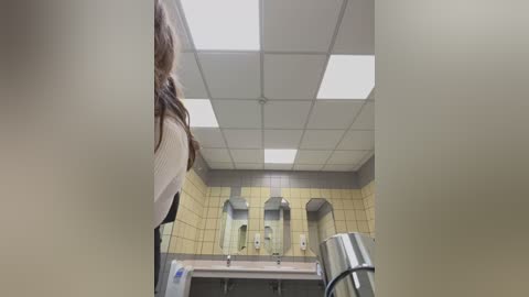 Media: Video of a bathroom stall with a woman in a white sweater standing, tiles, and a ceiling with fluorescent lights.