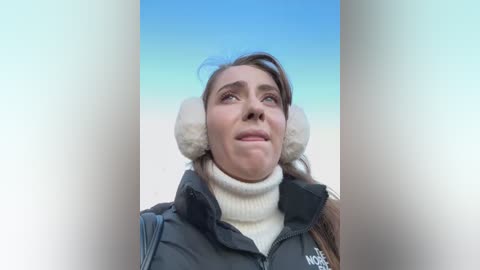 Media: Video of a Caucasian woman with fair skin, wearing a black puffy jacket, beige turtleneck, and white earmuffs, gazing upward against a clear blue sky.