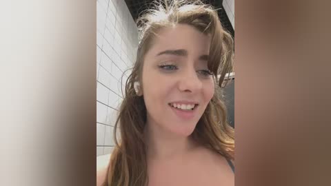 Media: Video of a young Caucasian woman with light brown hair, smiling, in a tiled bathroom. She has light skin, no makeup, and is topless, wearing a white bra.