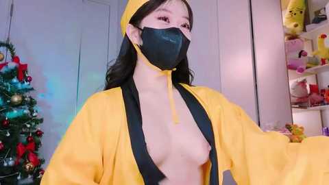 Media: Video of an Asian woman with long black hair, wearing a yellow graduation gown, black face mask, and revealing black bra, standing in a brightly lit room with a Christmas tree and stuffed toys in the background.