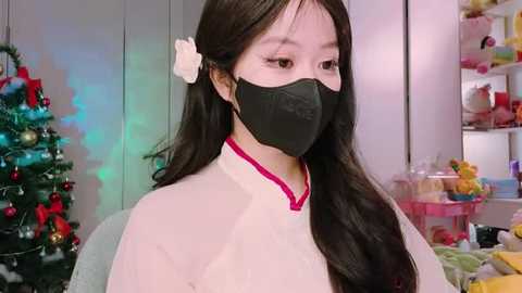 Media: Video of an East Asian woman with long black hair, wearing a white dress, black face mask, and a white flower in her hair. Background features a Christmas tree, toys, and colorful decorations.