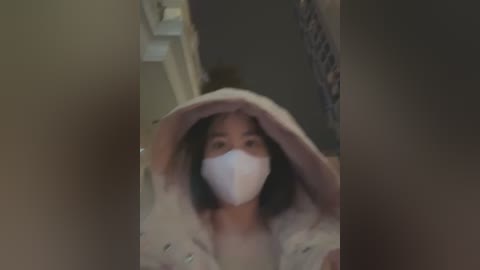 Media: A blurry, dark video of a woman wearing a white face mask, black hair, and a beige coat, standing indoors with a hand on her head.