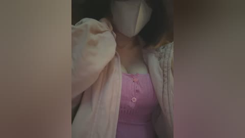 Media: Video of a person with a pale complexion, wearing a pink button-up blouse, a beige cardigan, and a white mask, positioned in a dimly lit room with a blurred background.