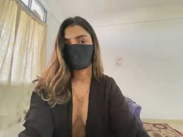 Media: Video of a light-skinned woman with long, wavy, dark-brown hair, wearing a black mask and a black open jacket, standing in a plain room with beige curtains and a patterned rug.