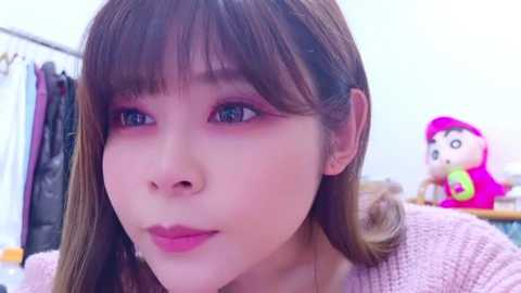 Media: Video of a young East Asian woman with light skin, straight brown hair, and blue eyes, wearing a pink sweater, looking slightly to the left. Background features a pink plush toy and hanging clothes.