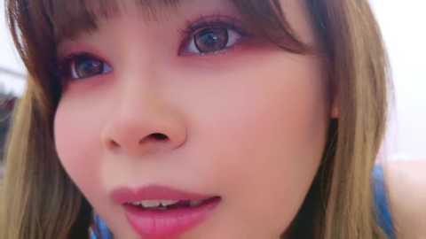 Media: Close-up video of a young Asian woman with fair skin, light brown hair, and blue eyes. She has a slight smile, wearing light makeup and a blue top.